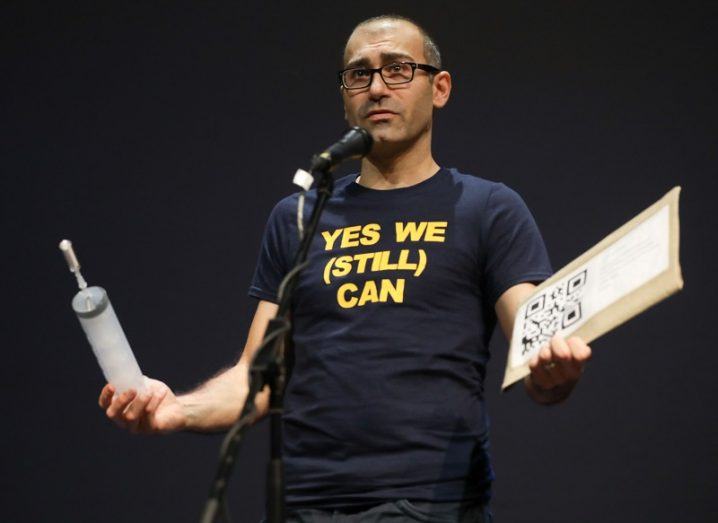 Dr Luca Mirimin holding props on stage while speaking into a microphone against a black background.
